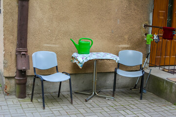 A simple outdoor seating area with two chairs and a round table, a green watering can, set against a rustic cream-colored wall.