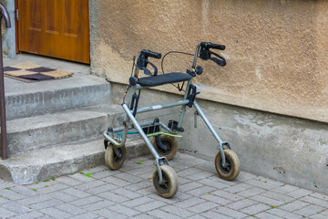 A silver walking frame with wheels, positioned on grey concrete steps leading up to a doorway,...