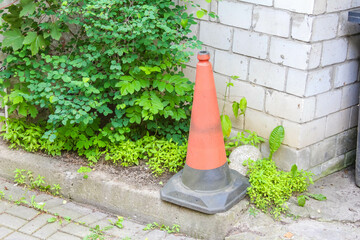 A bright orange traffic cone placed on a grey concrete surface beside a brick wall and green...