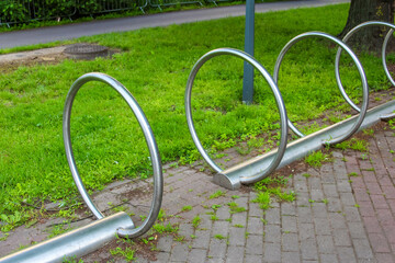 A row of modern, minimalist bicycle racks in a public park area with green grass and paved paths under a bright sky.