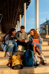 Three cheerful university students are sitting on campus stairs, sharing content on a smartphone,...