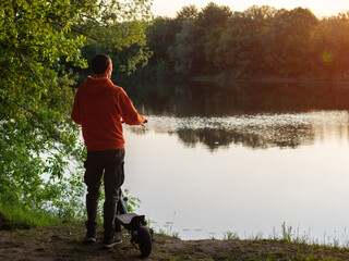 A man on an electric scooter stands by the river and enjoys nature at sunset.