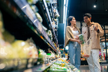 Naklejka na ściany i meble Happy Asian family couple using reusable cotton shopping bag buying food at supermarket. Man and woman enjoy sustainable living and healthy lifestyle choosing organic vegetable and fruit on shelf.