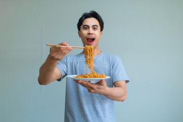 Happy young Asian man enjoying a plate of noodles with chopsticks, showing an excited expression on a clean background, perfect for food, culture, or lifestyle themes