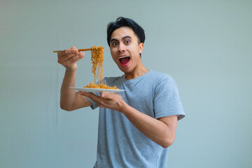 Happy young Asian man enjoying a plate of noodles with chopsticks, showing an excited expression on a clean background, perfect for food, culture, or lifestyle themes