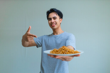 A happy young Asian man from Indonesia holds and points to a plate of noodles with excitement on a clear background, showcasing delicious food and joyful mood