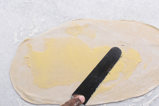 Overhead view of butter being spread on rolled out paratha dough, top view of flaky flatbread dough being covered with ghee, process of making paratha	