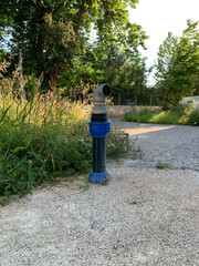 Fire hydrant on concrete pad, made of metal and plastic, with curved spigot for fire hose connection, grass, flowers around, trees, bushes, fence, technical infrastructure and nature in background