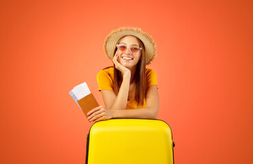 Happy young woman in sunglasses holding passport and tickets, leaning on big yellow suitcase, ready to travel. Cheerful lady traveller posing over yellow studio background, wearing summer hat © Prostock-studio
