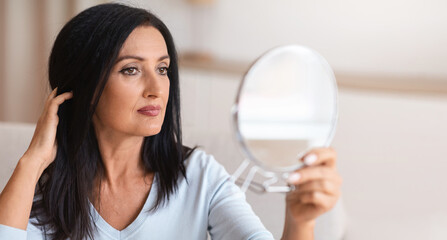Portrait of beautiful middle-aged brunette lady holding mirror and looking at her reflection, touching her hair with hand. Confident adult woman thinking about skin care and body treatment