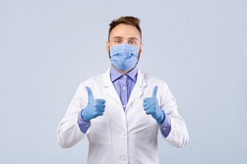 Millennial male doctor in uniform, face mask and gloves looking at camera and showing thumbs up gesture over grey studio background. Physician wearing protection during coronavirus outbreak