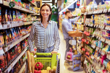 Portrait Of Smiling Woman With Shopping Cart In Supermarket Buying Groceries Food Walking Along The Aisle And Shelves In Grocery Store. Female Buyer Choosing Healthy Products In Mall
