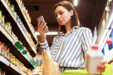 Modern Shopping. Young Lady Using Phone Buying Food Groceries Standing Wih Trolley Cart In Supermarket. Woman With Smartphone Putting Bottle In Basket, Reading Checklist, Cheking Sales Online