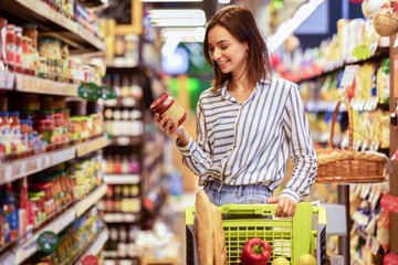 Portrait Of Smiling Woman With Shopping Cart In Supermarket Buying Groceries Food Walking Along The Aisle And Shelves In Grocery Store, Holding Glass Jar Of Sauce, Choosing Healthy Products In Mall