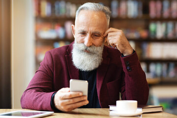 Senior grey-haired man with long beard drinking coffee at cafe, using smartphone, planning his day,...
