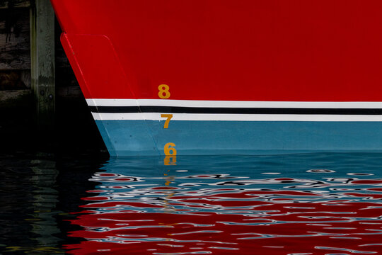 A large red, white, black and teal blue ship with waterline marks painted with yellow paint. The numbers reflect the line where the hull meets the surface of water. The water has ripples on the surfac