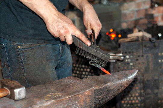 A forger wearing blue jeans and a t-shirt holds fire thongs and a metal twisted bolt on a vintage anvil. There's a rounding hammer tool and a wire brush in the blacksmith foundry shop with a furnace. - Powered by Adobe