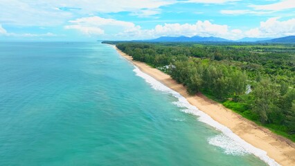 The drone captures a mesmerizing stretch of beach, where the smooth, turquoise water meets golden sands, framed by lush trees a perfect combination of water, land, and greenery. Phuket, Thailand.
