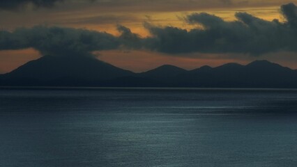 A captivating aerial view of dark, dramatic mountains silhouetted against the fading light of a colorful sunset, with calm waters reflecting the serene yet mysterious atmosphere. Phuket, Thailand.
