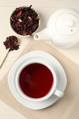 Delicious hibiscus tea in cup, teapot, dry roselle sepals and spoon on light wooden table, flat lay