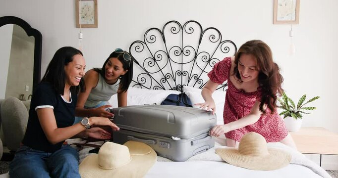 Diverse female friends opening suitcase on bed, sorting hats before checking trip plan on tablet