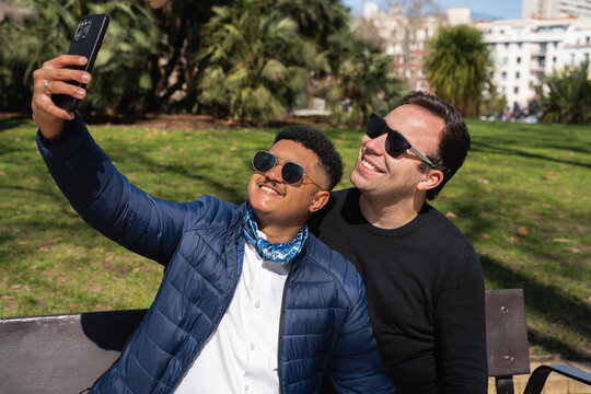 Two smiling men take a selfie with a phone in a city park