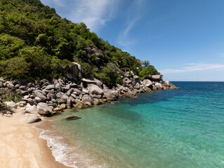 Tropical shoreline with rocky formations, lush greenery, and blue sea under sunny skies. Thailand, Koh Tao.