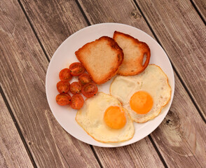 A plate of delicious breakfast, two fried eggs with cherry tomatoes and two wheat toasts on a wooden table.