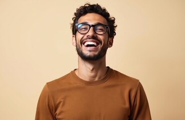 Young Brazilian man with glasses laughs on beige background. Happy guy with beard, trendy hairstyle, and smile. Positive emotions, joy, fun. Latin model person smiling showing teeth.