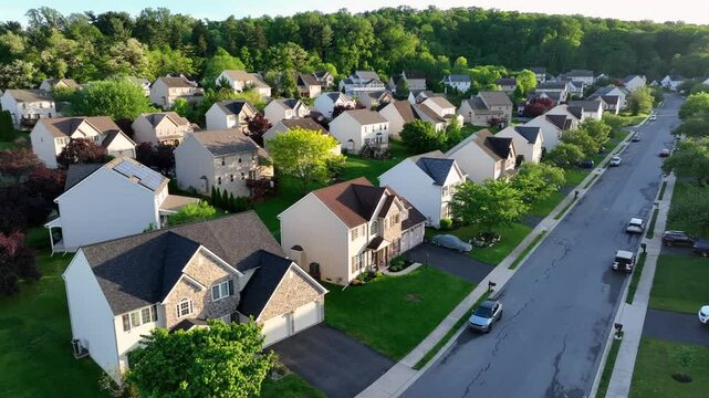 Classical and brick style two-story homes with Solar panel on roof. Located on hill with green trees at sunset. Aerial approaching wide shot. Cars on street. Small american neighborhood of PA.