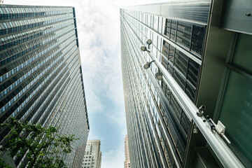 Two sleek skyscrapers stretch into the sky between narrow streets in Midtown Manhattan. A streetlight and security cameras are visible in the foreground, adding a sense of urban infrastructure.