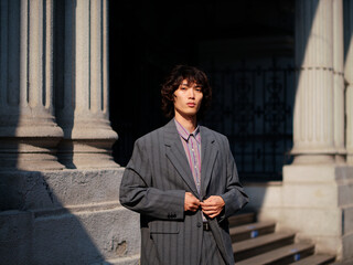 Portrait of handsome Chinese young man wearing gray suit posing in the street, young guy with black curly hair with urban background. Male fashion, cool Asian young man lifestyle.