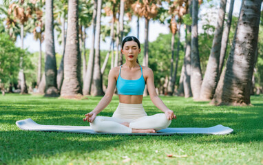 Asian woman meditating in a peaceful park setting, sitting cross-legged on green grass with trees in the background on a sunny day. Concept of mindfulness, self-care, and outdoor relaxation
