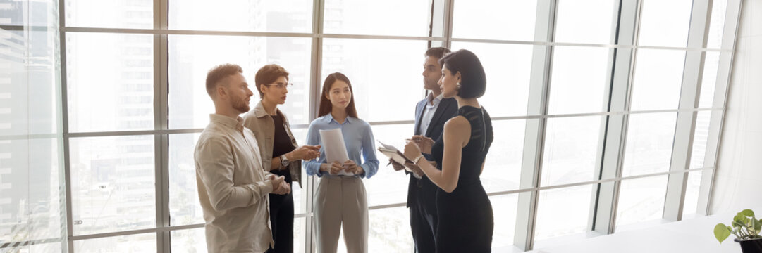 Group of five processionals discussing work-related matters gathered in modern office standing against panoramic window with skyscraper city view, wide copy-space image. Business interaction, teamwork