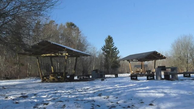 Calming scene of hiking trail vacation log and wooden picnic tables and grilling spot in winter nature during a gorgeous sunny day. The structures are modern and leafless bare trees surround them.