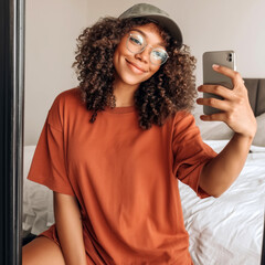 Women burnt orange oversized t-shirt mockup. Smiling black female model in sunglasses, hat, summer outfit, takes selfie in room. Casual short sleeve shirt mockup, indoors. Oversized crewneck tee mock