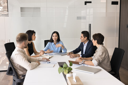 Professional team take part in meeting in office boardroom. Five businesspeople sit around conference table, engaged in discussion, brainstorming, strategizing, or reviewing important business topics