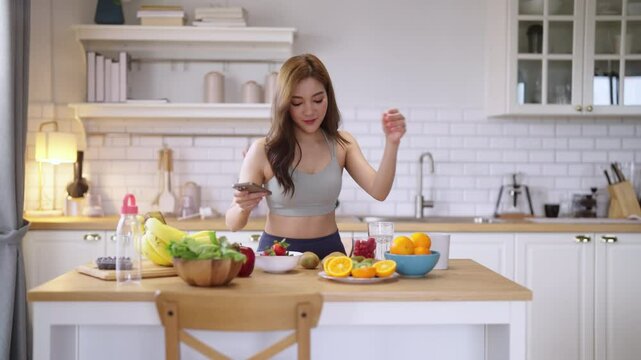Healthy Asian woman in activewear prepares a fresh fruit drink, slicing fruit and blending a protein shake in her modern pantry kitchen at home, promoting wellness and fit lifestyle.