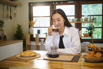 Asian female relaxing at a wooden kitchen counter, sipping a warm drink with a calm and peaceful expression.