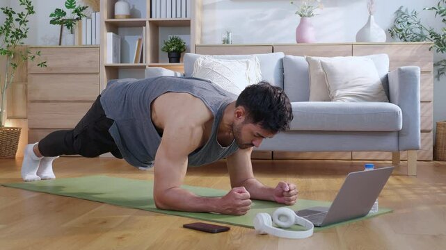 Young asian man doing plank exercise in living room at home while watching online workout tutorial on laptop with endurance and motivation, man training plank on yoga mat, digital fitness lifestyle.