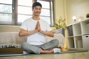 Calm Asian man meditating on mat, practicing mindfulness in a cozy living room.