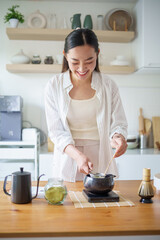 Young woman in casual outfit preparing matcha tea in home kitchen with traditional tools and...