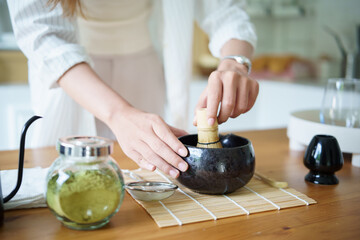 Close up of woman whisking matcha tea in a traditional black bowl on a wooden table.