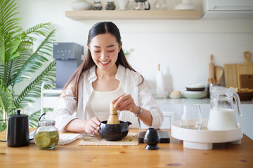 Young woman making matcha at home, surrounded by ingredients and utensils on a wooden table.