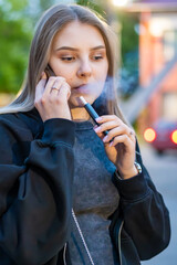 Young woman smoking e-cigarette talking on the phone in the street. Addiction concept. High quality...
