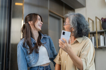 Asian senior mother and daughter smiling and talking using smartphone browsing social media, shopping online, watching movies or series, enjoying entertainment digital connection and bonding at home