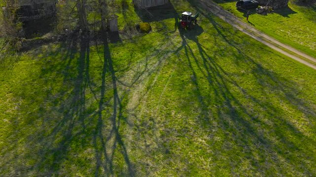 Aerial drone footage following a small retro and vintage tractor working in a sunny green grassy field during summer time at a countryside garden or a yard that has large trees that cast shadows on it