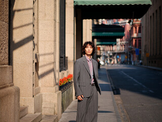 Portrait of handsome Chinese young man wearing gray suit posing in the street, young guy with black curly hair with urban background. Male fashion, cool Asian young man lifestyle.