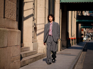 Portrait of handsome Chinese young man wearing gray suit walking in the street, young guy with black curly hair with urban background. Male fashion, cool Asian young man lifestyle.