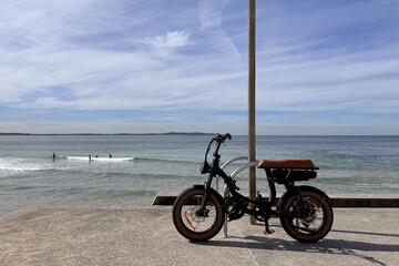 Obraz premium e-bike, fat tyre electric bicycle tied to post outdoors at the beach on a sunny day with distant view of people swimming in water